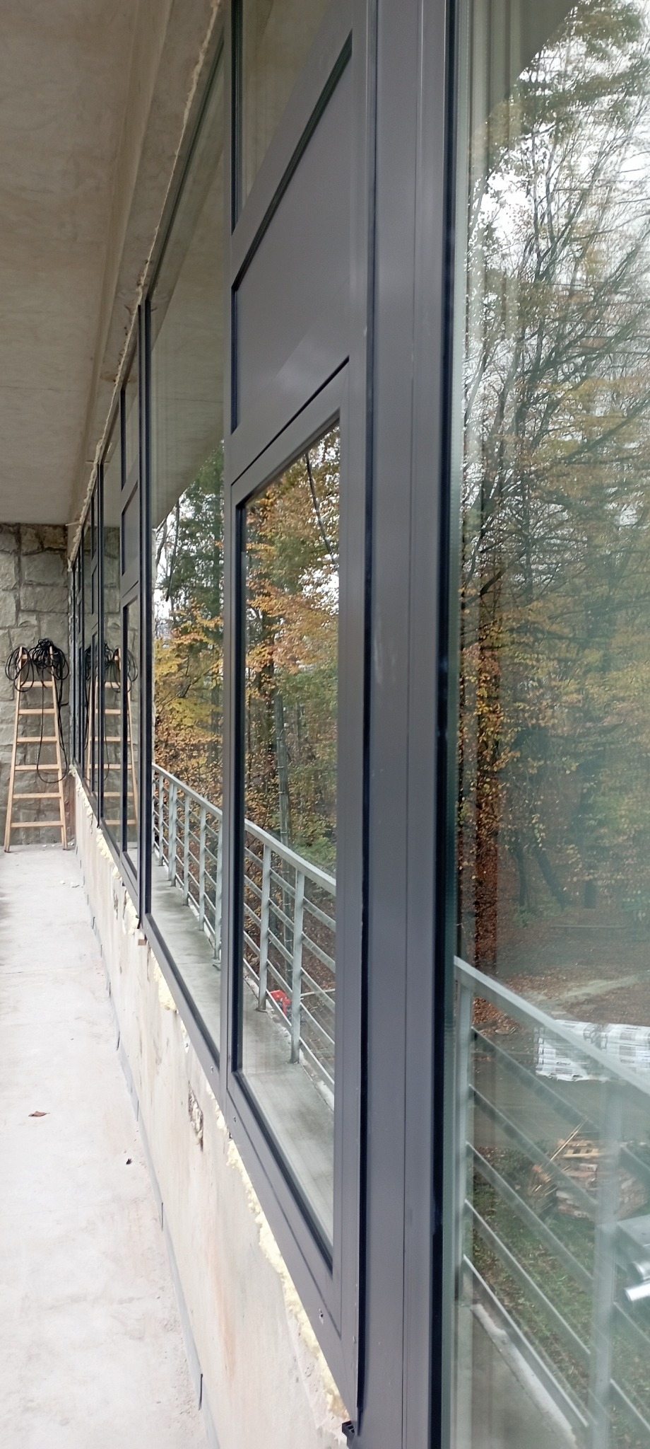Montaż okien z szarą ramą na balkonie, widoczna drabina i jesienny krajobraz za oknem.