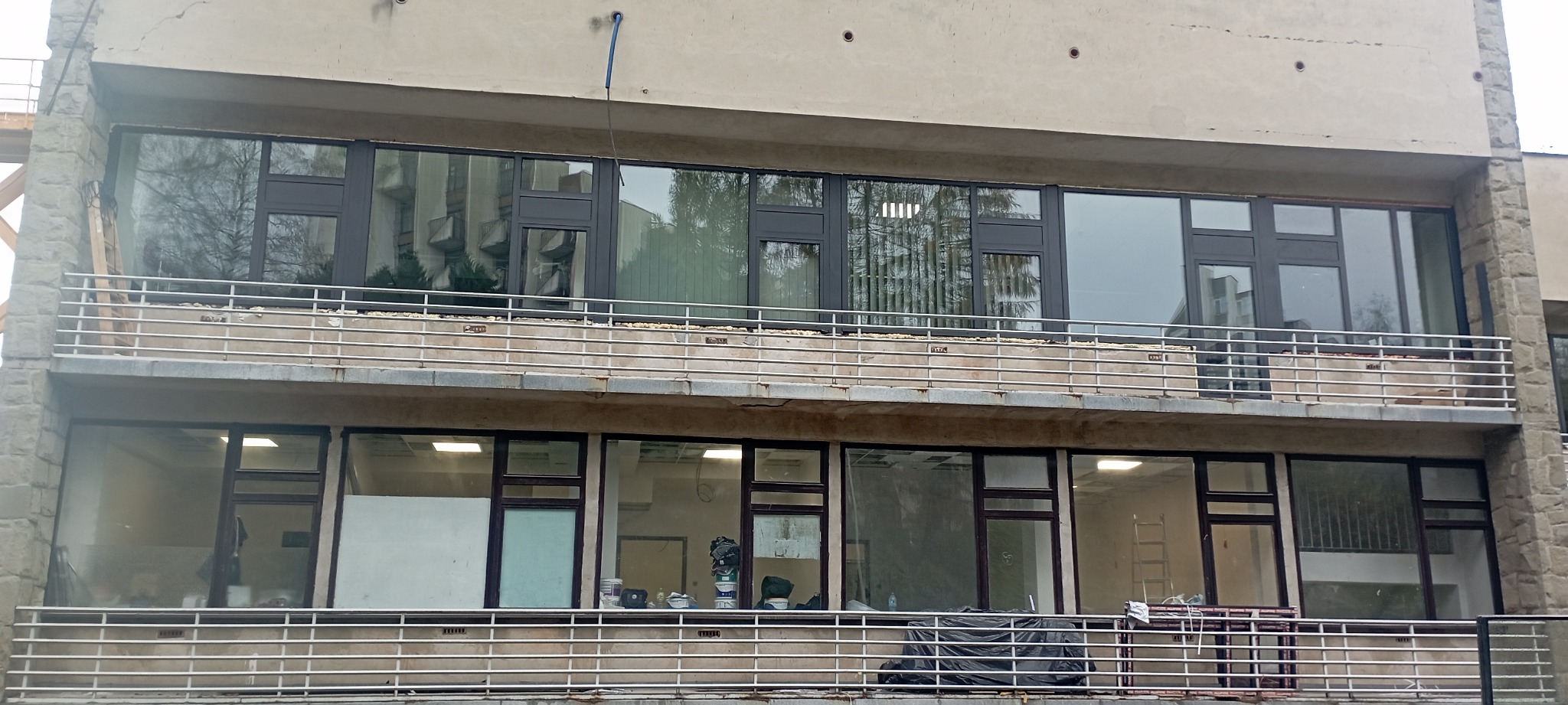 Budynek z zamontowanymi oknami w ciemnych ramach, widoczne prace wykończeniowe wewnątrz pomieszczeń, balkon z metalową barierką.