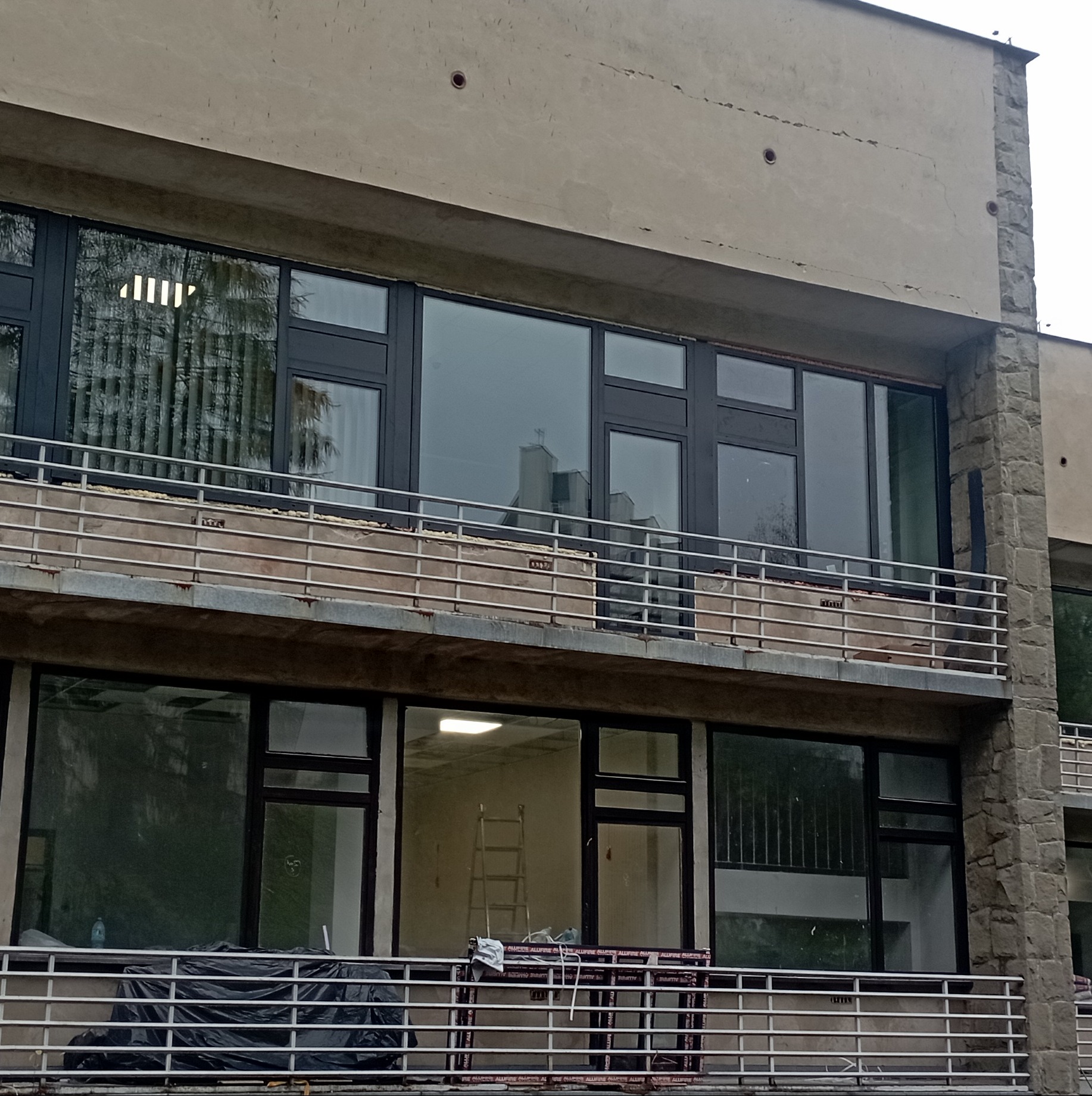 Budynek w trakcie remontu z zamontowanymi oknami z czarnymi ramami, widoczne rusztowania i drabina wewnątrz pomieszczenia.