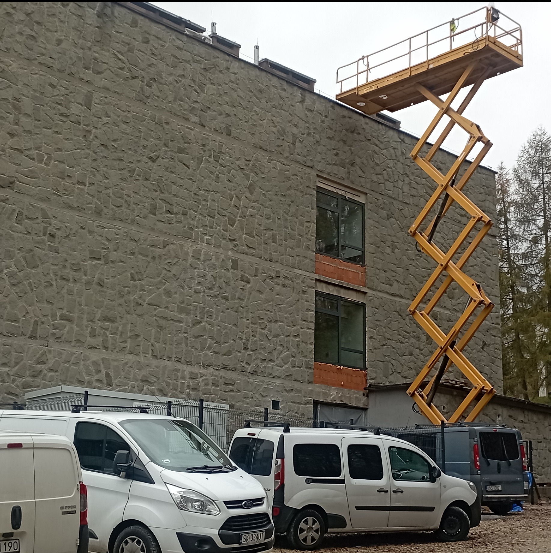 Żółty podnośnik koszowy ustawiony przy budynku z kamienną elewacją, widoczne okna z ceglanym obramowaniem i zaparkowane busy.