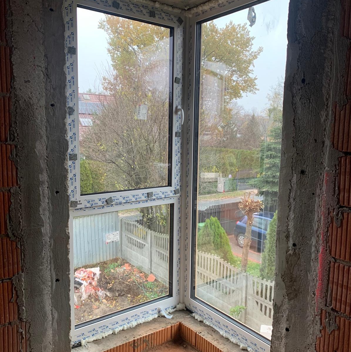 Narożne okno z białymi ramami, widok z surowego wnętrza na ogród w Warszawie, z widocznymi śladami pianki montażowej.