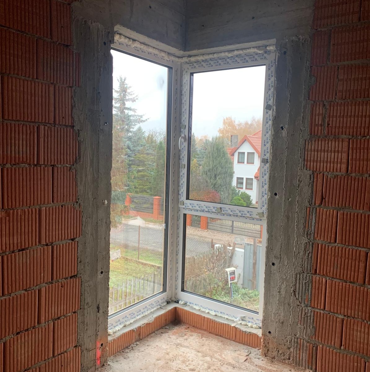 Narożne okno z białymi ramami zamontowane w surowym murze z czerwonej cegły, widok na dom i drzewa za oknem.