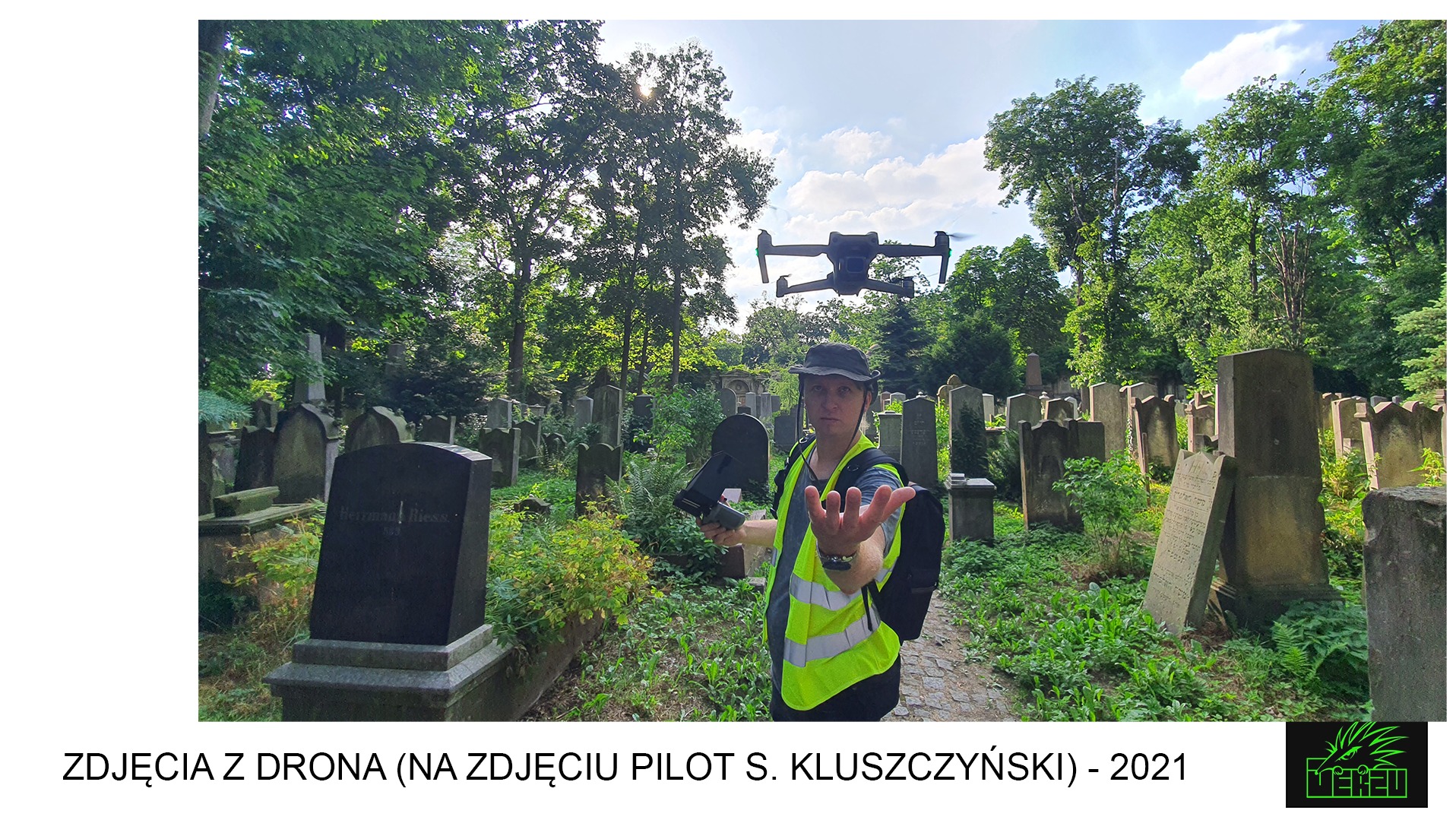 Pilot drona w kamizelce odblaskowej na tle cmentarza, dron w powietrzu, kontroler w ręku.