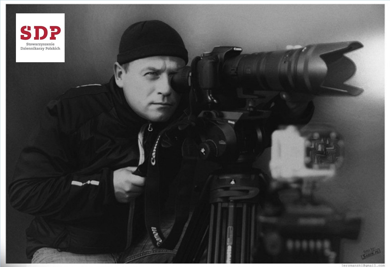 Czarno-białe zdjęcie mężczyzny w czapce, patrzącego przez wizjer profesjonalnej kamery wideo na statywie, z logo SDP w lewym górnym rogu.