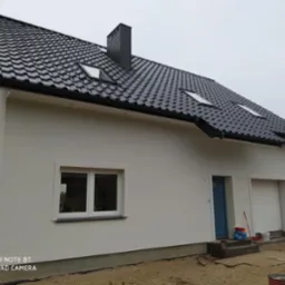 Nowoczesny dom z czarną dachówką i białymi ścianami, widok z boku. Elementy wykończeniowe, okna z białymi ramami i garaż. Dom w trakcie prac wykończeniowych.