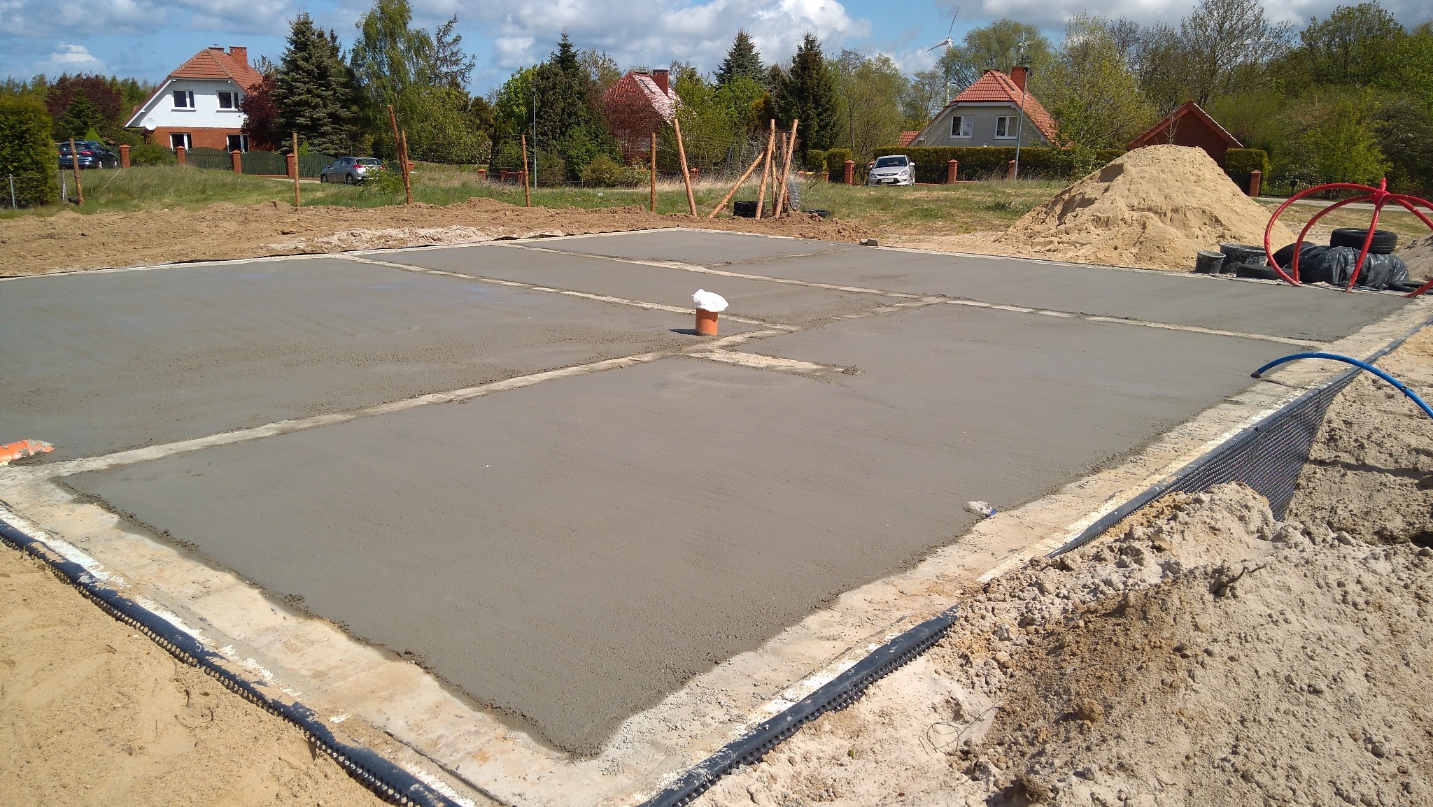 Wylana płyta fundamentowa z widoczną izolacją pionową, przygotowana do dalszych prac budowlanych. Na powierzchni rury i biała folia. W tle zabudowa jednorodzinna.