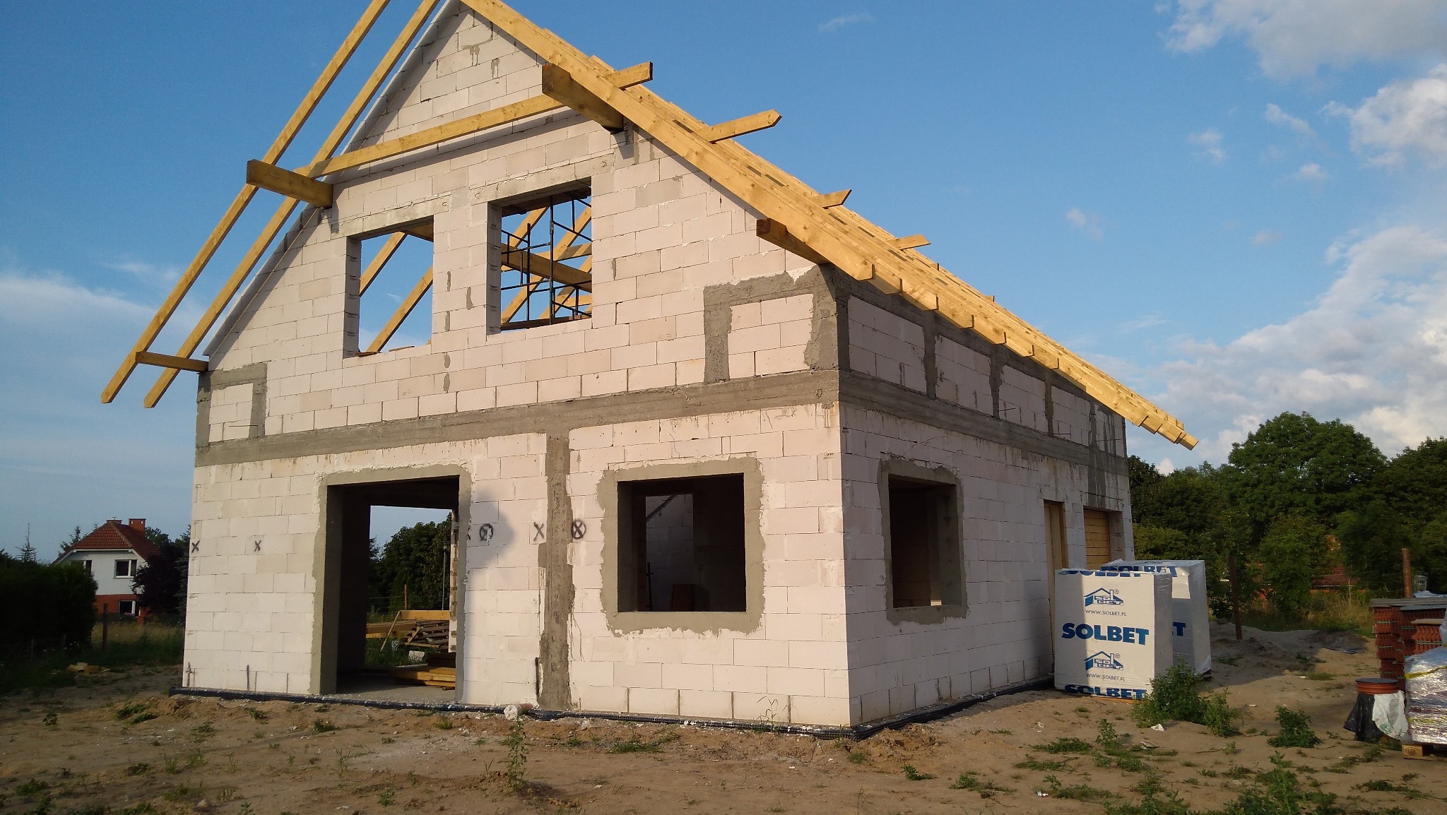 Niedokończony dom z białych bloczków Solbet, widoczne drewniane elementy konstrukcji dachu, okna bez szyb, w tle zieleń i błękitne niebo.