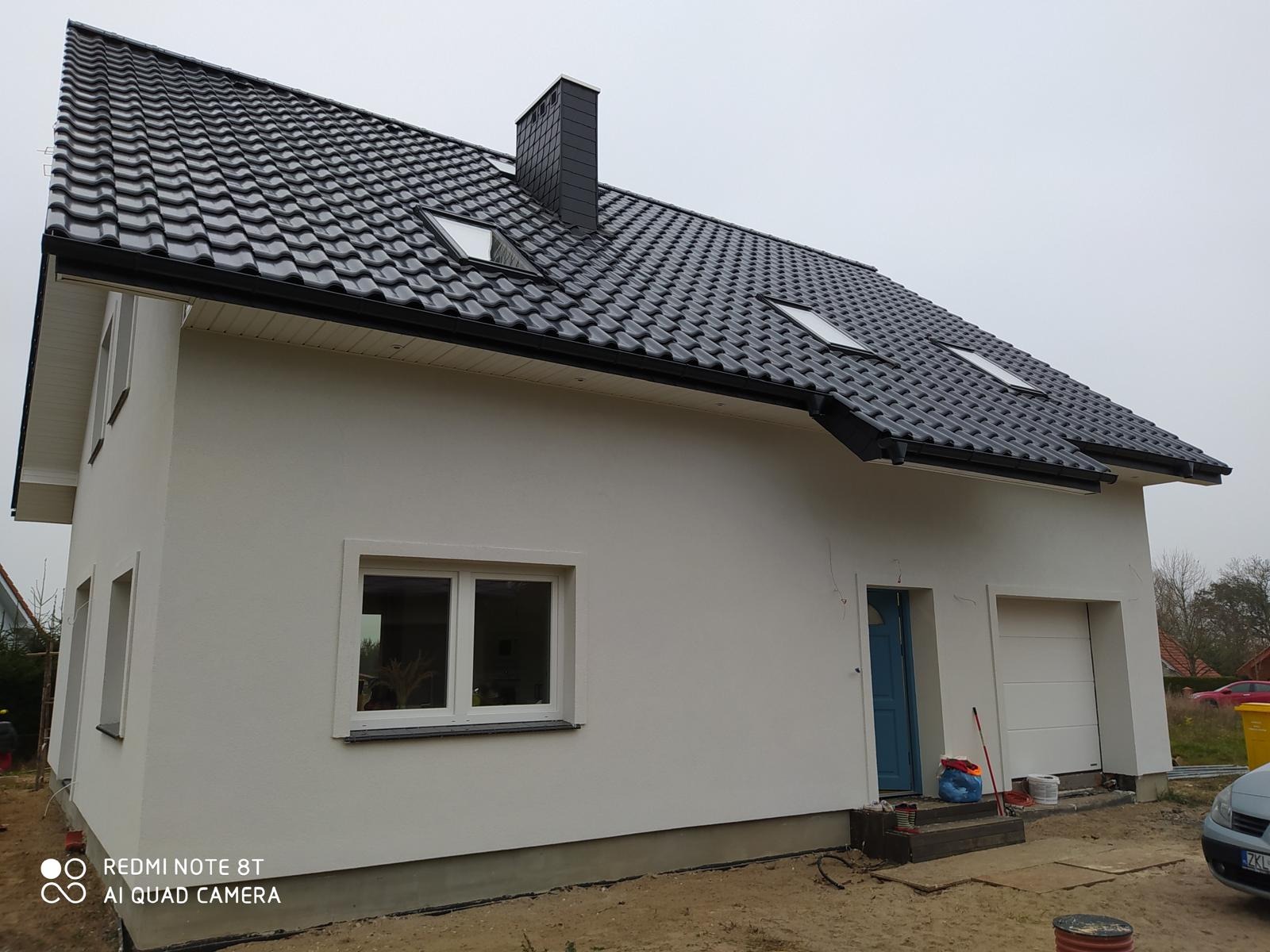 Nowoczesny dom z czarną dachówką i białymi ścianami, widok z boku. Elementy wykończeniowe, okna z białymi ramami i garaż. Dom w trakcie prac wykończeniowych.