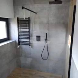 Nowoczesna kabina prysznicowa walk-in z drewnopodobną podłogą, szarymi płytkami ściennymi, czarną armaturą i grzejnikiem drabinkowym.