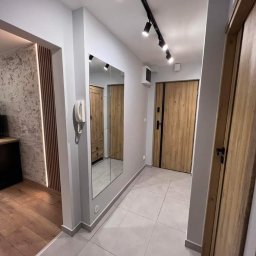 Manbud - Nowoczesny przedpokój z szarymi płytkami podłogowymi, drewnianymi drzwiami i lustrem odbijającym szafę. Czarny system oświetlenia szynowego na suficie.