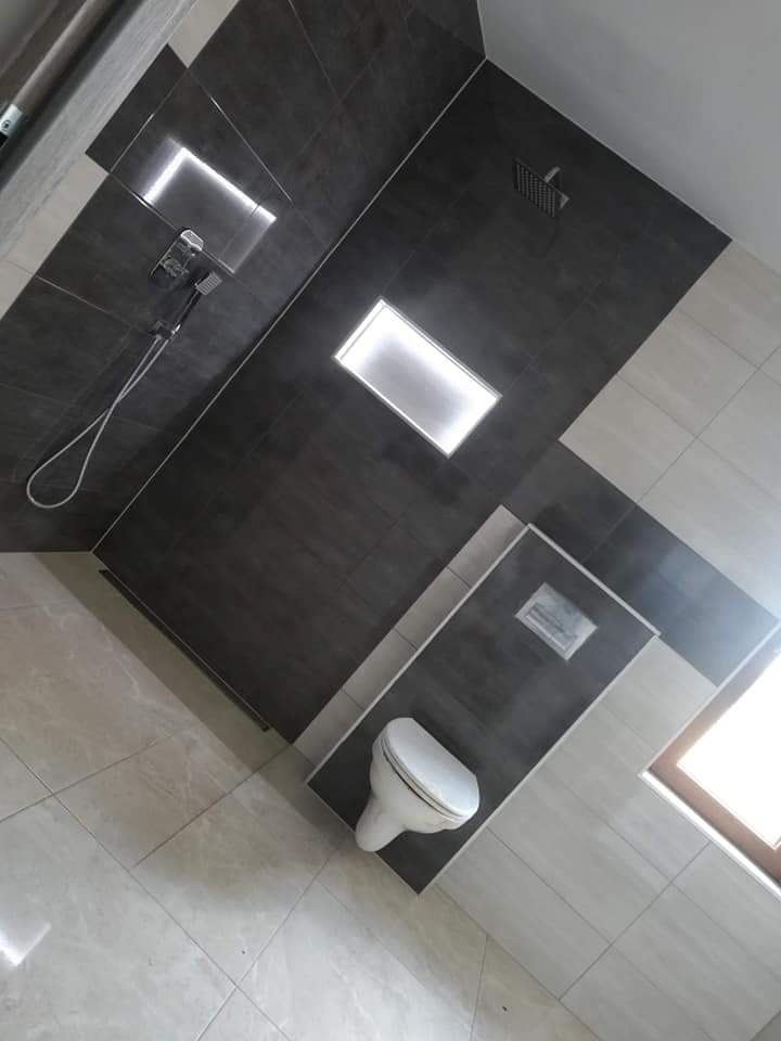 Nowoczesna łazienka z ciemnymi płytkami na ścianach, podświetlanymi wnękami, jasnymi płytkami podłogowymi i wiszącą toaletą.