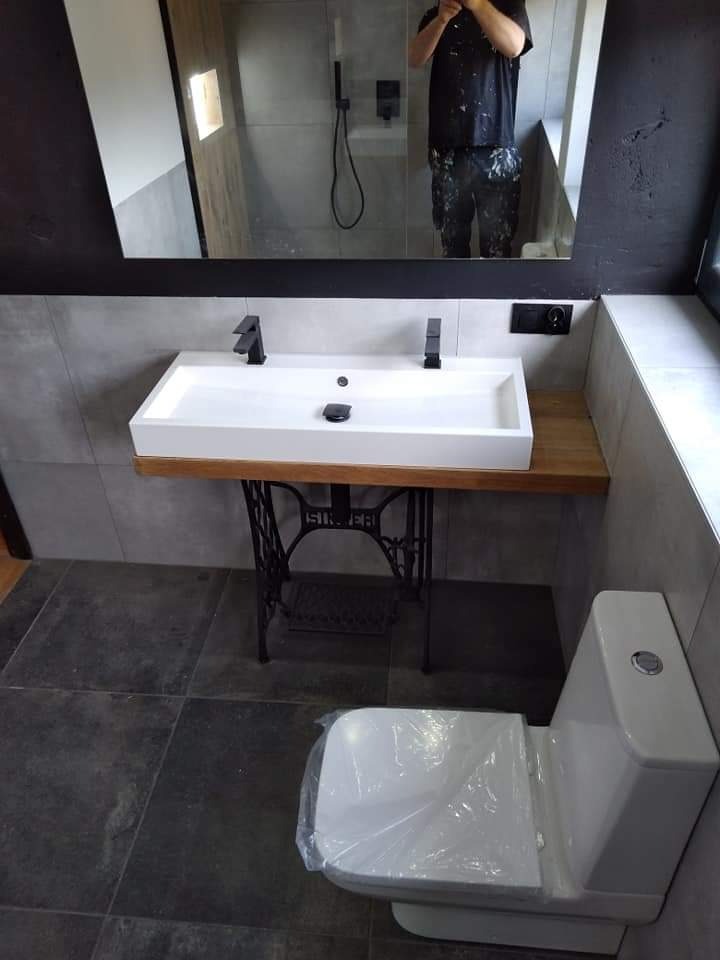 Nowoczesna łazienka z umywalką na blacie z drewnianym wykończeniem, wspartą na metalowej podstawie od maszyny do szycia Singer; w lustrze odbija się osoba w ubraniach roboczych z plamami farby...