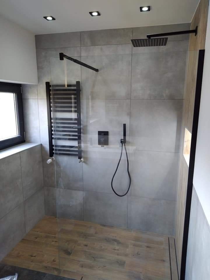 Nowoczesna kabina prysznicowa walk-in z drewnopodobną podłogą, szarymi płytkami ściennymi, czarną armaturą i grzejnikiem drabinkowym.