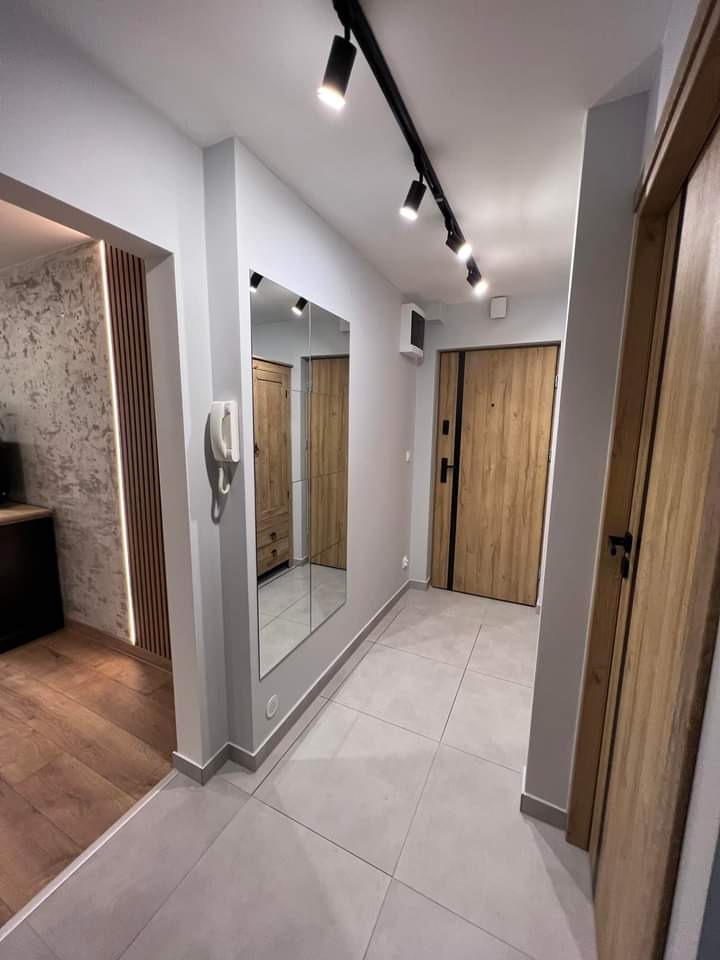 Nowoczesny przedpokój z szarymi płytkami podłogowymi, drewnianymi drzwiami i lustrem odbijającym szafę. Czarny system oświetlenia szynowego na suficie.