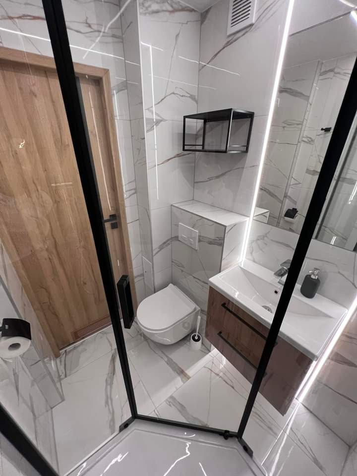 Nowoczesna łazienka z kabiną prysznicową typu walk-in, wykończona białymi płytkami imitującymi marmur z delikatnymi szarymi żyłami, drewnianą szafką pod umywalką i czarnymi akcentami w postaci ramy...