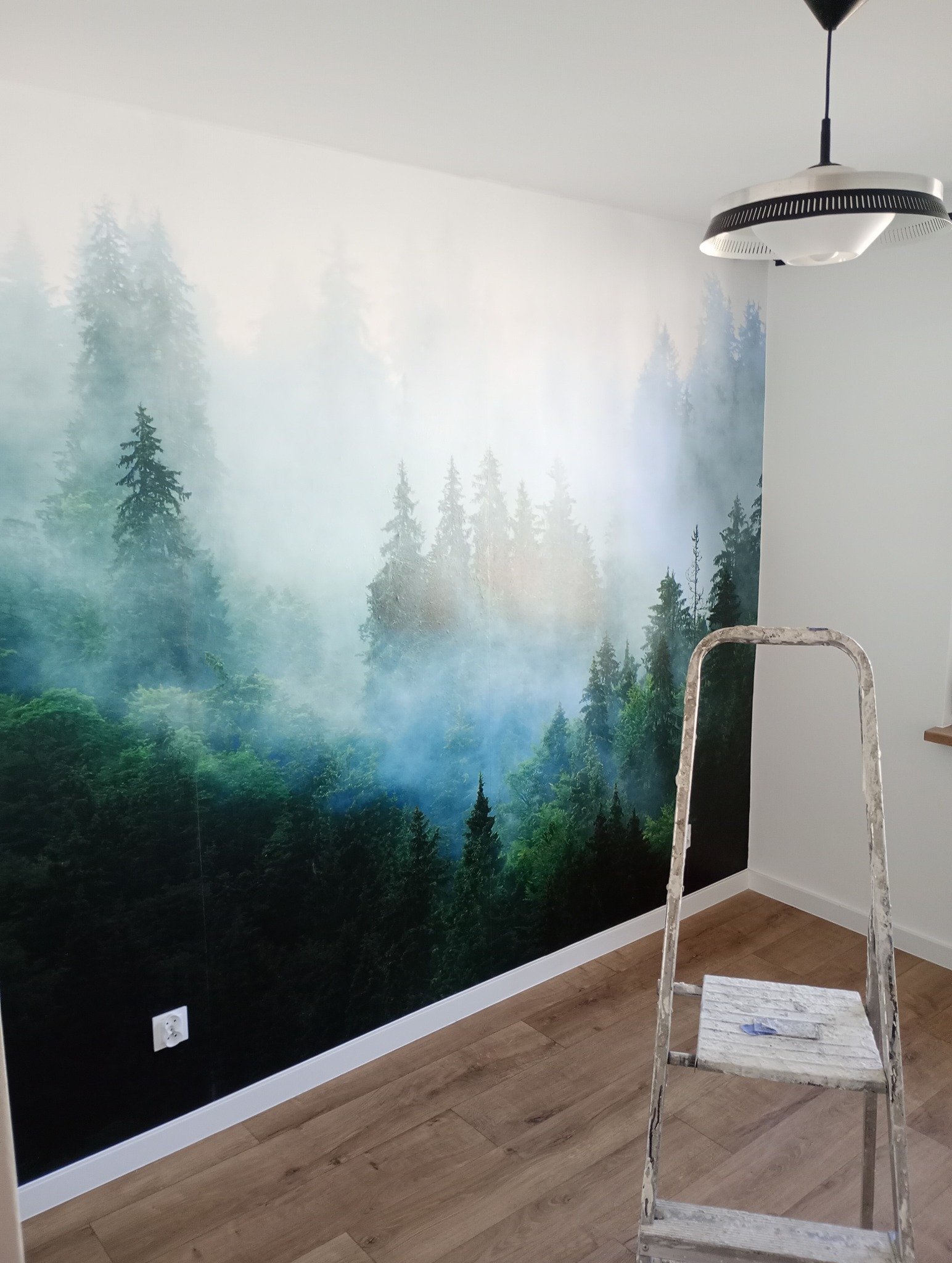 Ściana pokoju z fototapetą przedstawiającą mglisty las iglasty, widoczna drabina malarska i nowoczesna lampa sufitowa.