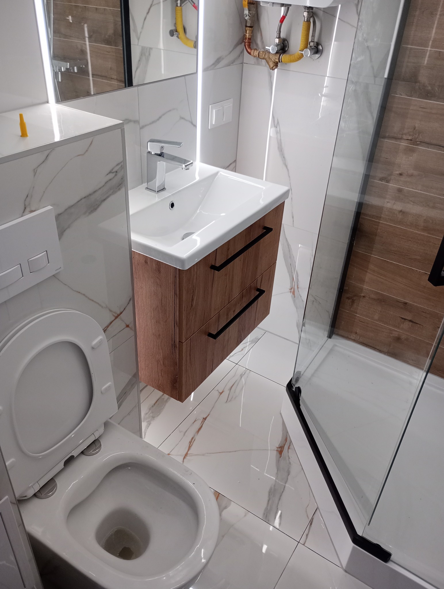 Nowoczesna łazienka po remoncie: białe płytki z marmurkowym wzorem, wisząca szafka pod umywalką z drewnopodobnymi frontami i czarnymi uchwytami, kabina prysznicowa ze szklanymi drzwiami i białą...