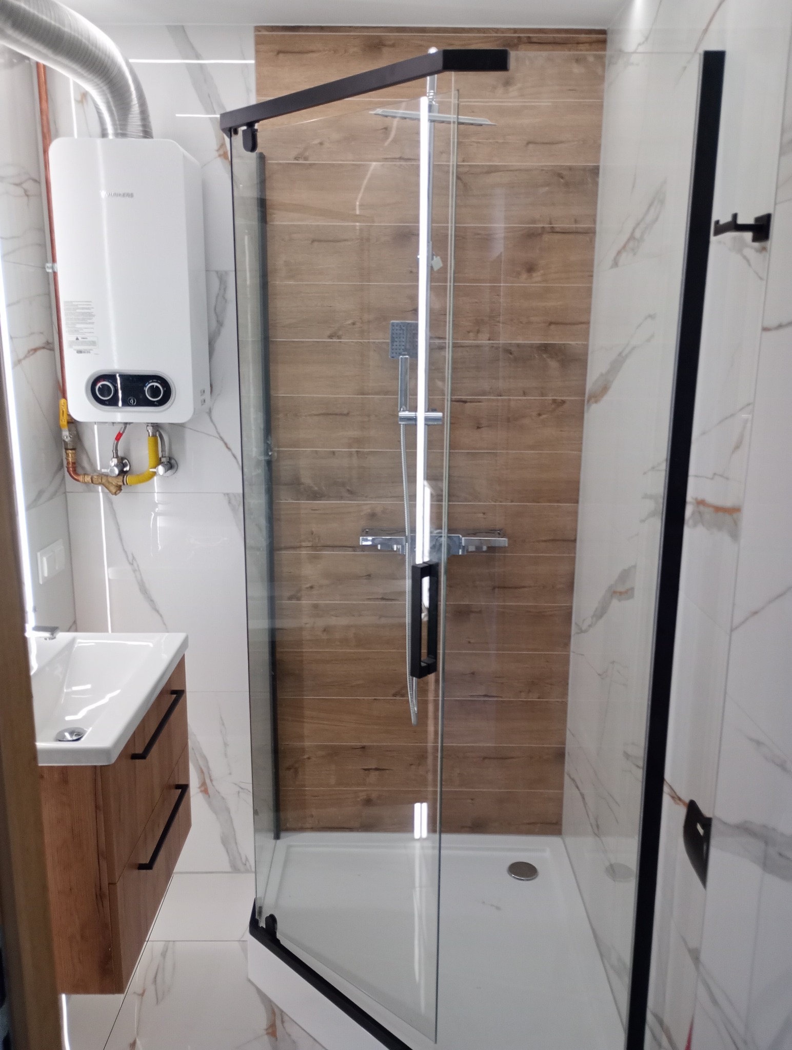 Nowoczesna kabina prysznicowa z drewnopodobnymi płytkami na ścianie, umywalka z drewnianą szafką i podgrzewacz wody w wykończonej łazience.