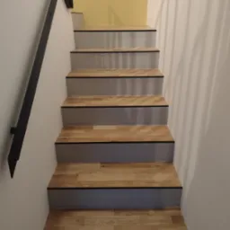 Schody drewniane z czarnymi krawędziami, szare stopnice, czarna metalowa poręcz, ściana pomalowana na biało i żółto w tle.