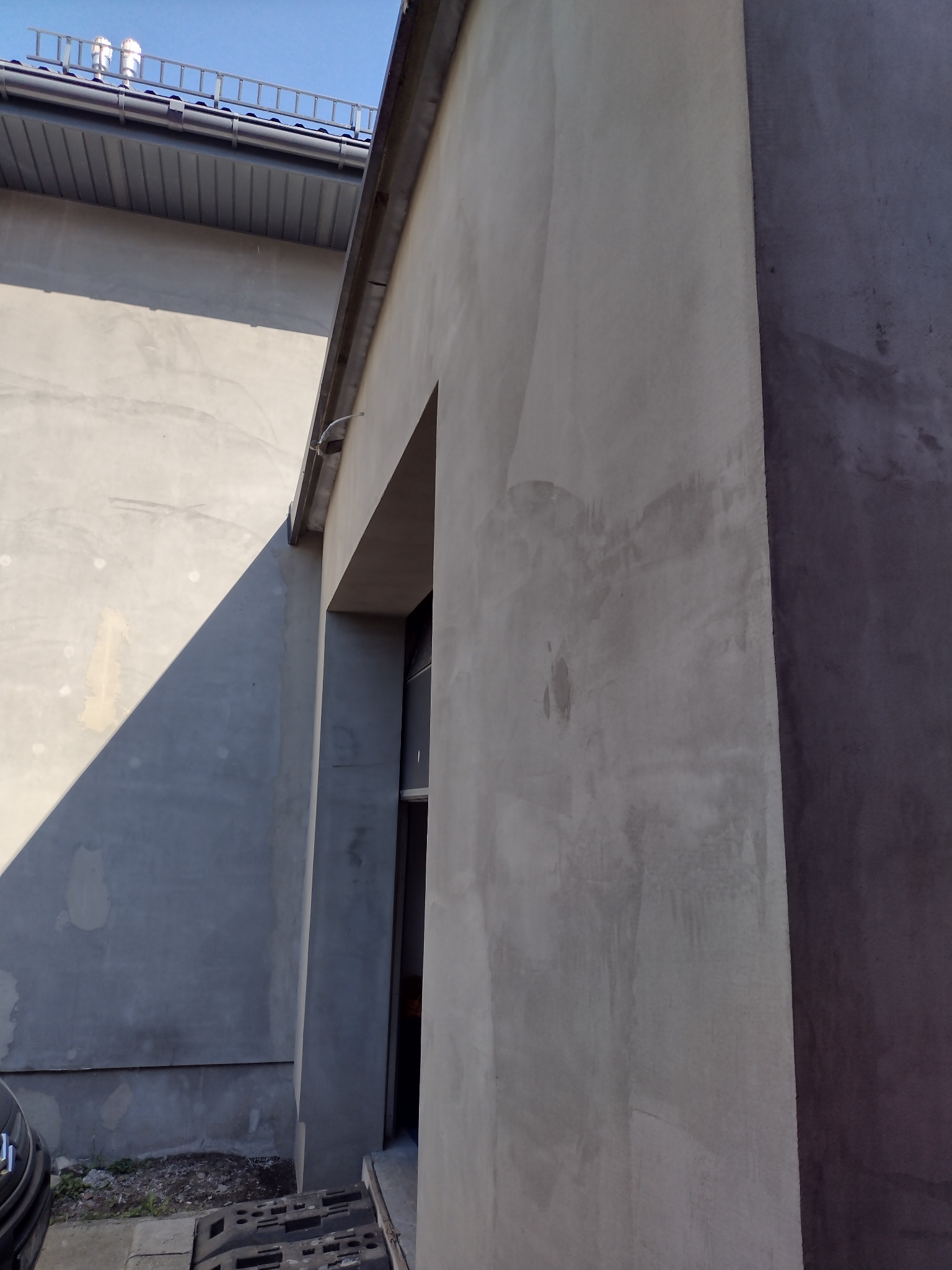 Surowa ściana budynku w trakcie tynkowania, widoczne nierówności i faktura, zarys okna/drzwi w głębi, fragment dachu z drabinką na górze.