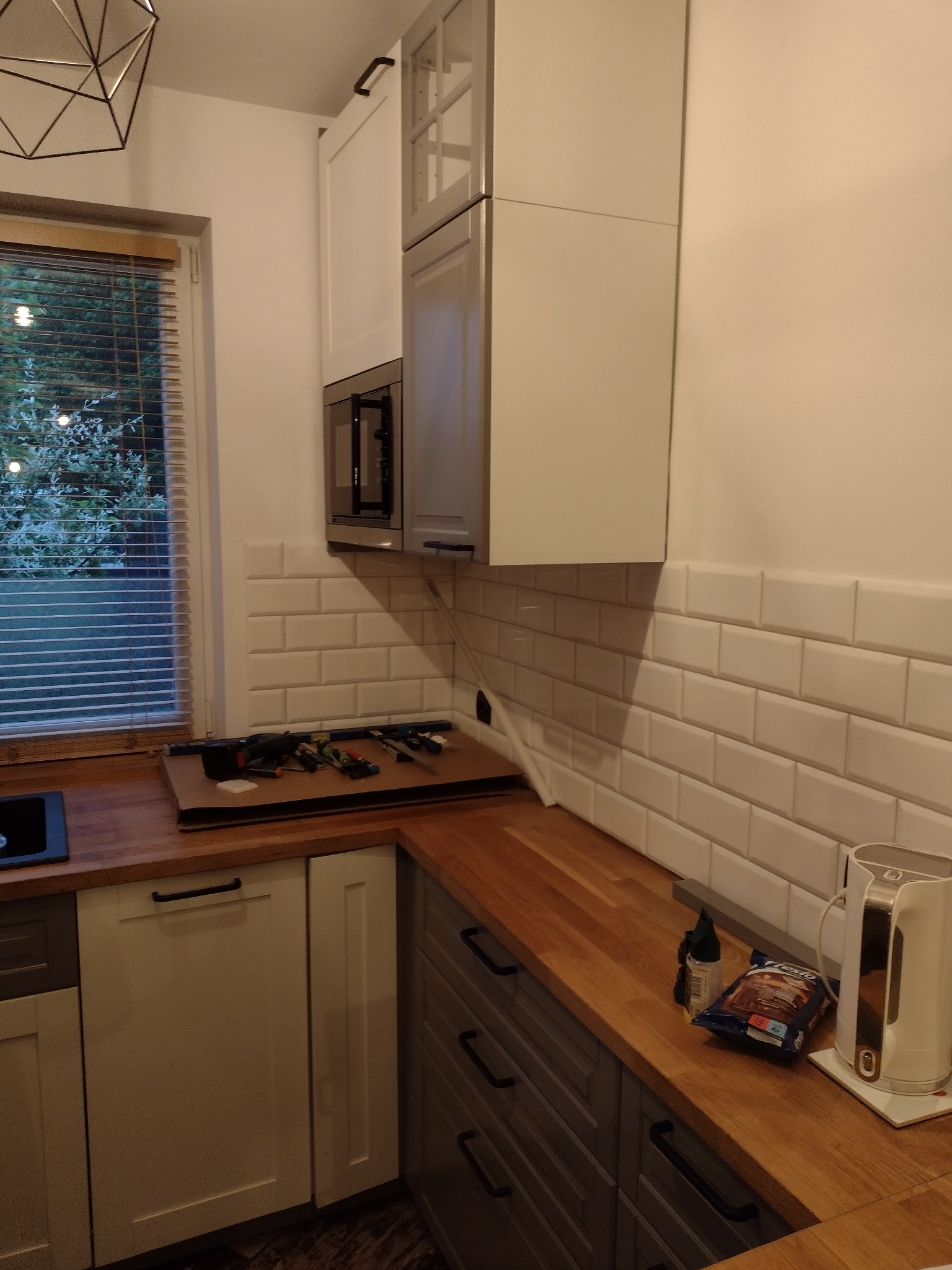Wnętrze kuchni w trakcie prac wykończeniowych: białe płytki ułożone na ścianie, drewniany blat, biało-szare szafki, okno z żaluzjami i narzędzia na blacie.