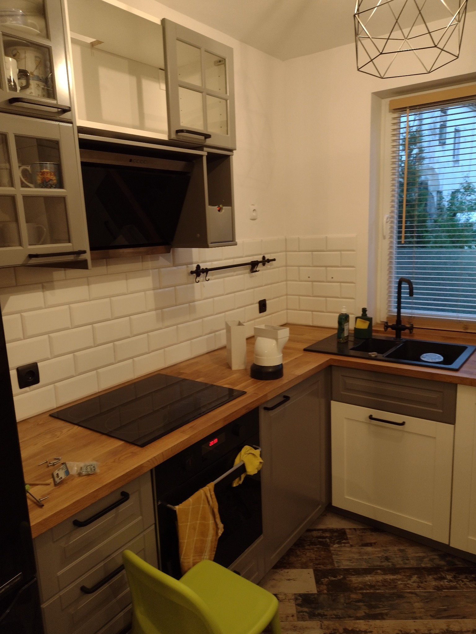 Szare szafki kuchenne z drewnianym blatem, czarną płytą indukcyjną i piekarnikiem, białe płytki na ścianie, czarny kran przy oknie z żaluzjami.