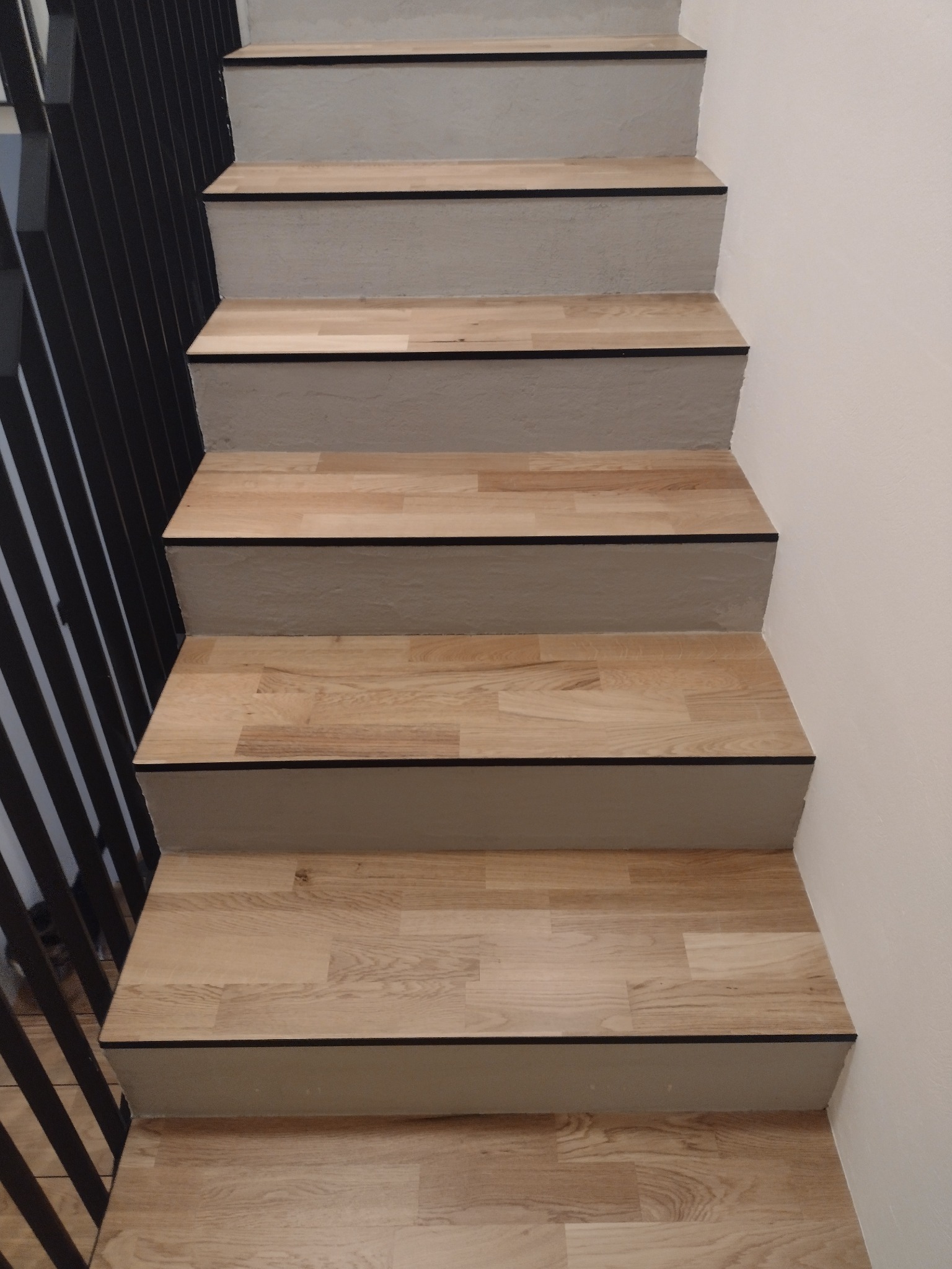 Widok z góry na drewniane schody z czarnymi krawędziami, z czarną metalową balustradą po lewej stronie i gładką, jasną ścianą po prawej.