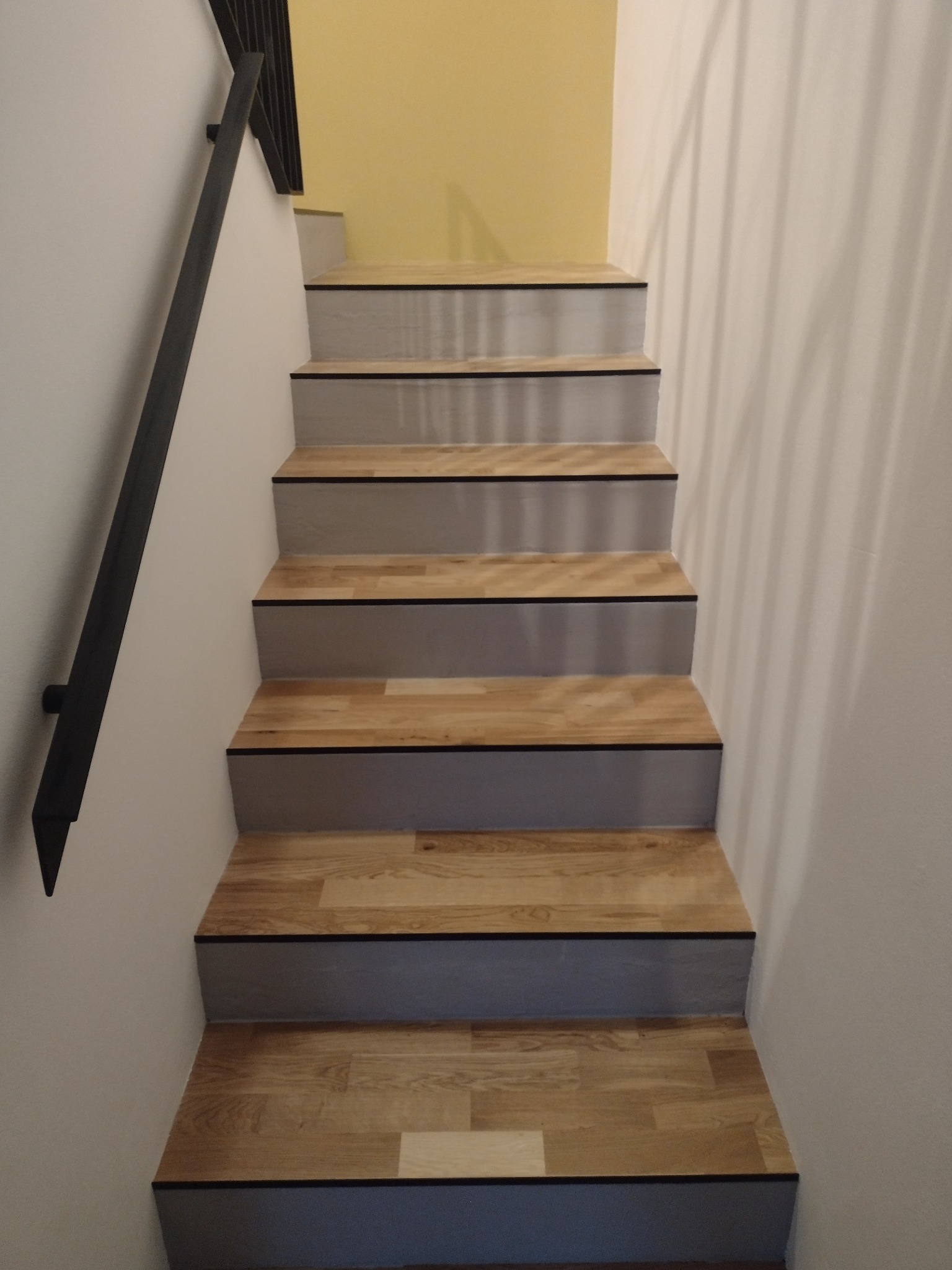 Schody drewniane z czarnymi krawędziami, szare stopnice, czarna metalowa poręcz, ściana pomalowana na biało i żółto w tle.
