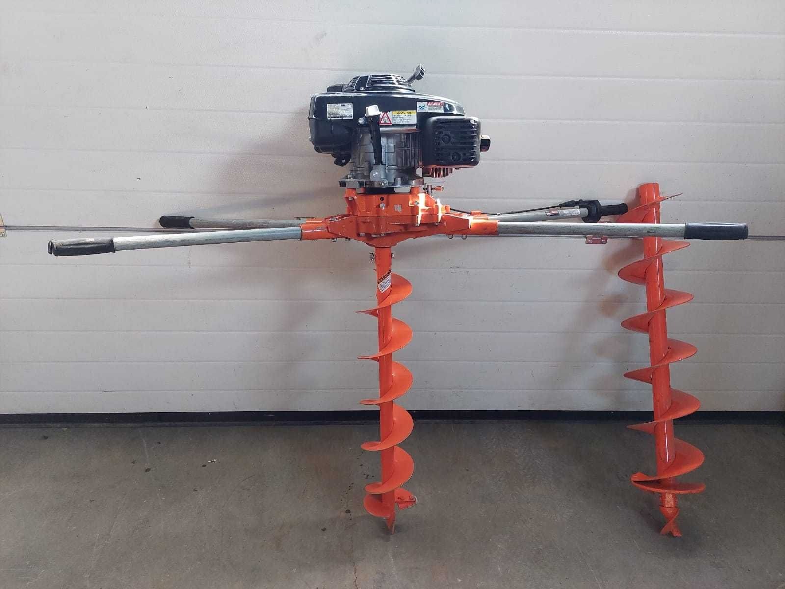 Dwuosobowa wiertnica glebowa z silnikiem spalinowym, pomarańczowa, z dwoma świdrami o spiralnym kształcie, na tle białej ściany garażowej.