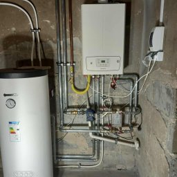 HYDRO-BLANEX Tomasz Żądło - Biały piec gazowy Immergas z podłączeniami, obok biały bojler Kospel. Widoczne rury miedziane i stalowe, zawory, żółty przewód gazowy. Surowe ściany w tle.