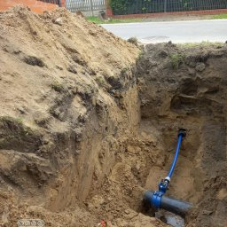 HYDRO-BLANEX Tomasz Żądło - Wykonany wykop z widocznym przyłączem wodociągowym z niebieską rurą, złączkami i zaworem. W tle ziemia, ulica i ogrodzenie posesji.