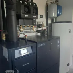 Czarny piec na paliwo stałe marki Kaman z podajnikiem, zbiornikiem i sterownikiem, obok biały zasobnik ciepłej wody. Widoczne rury i zawory instalacji grzewczej.