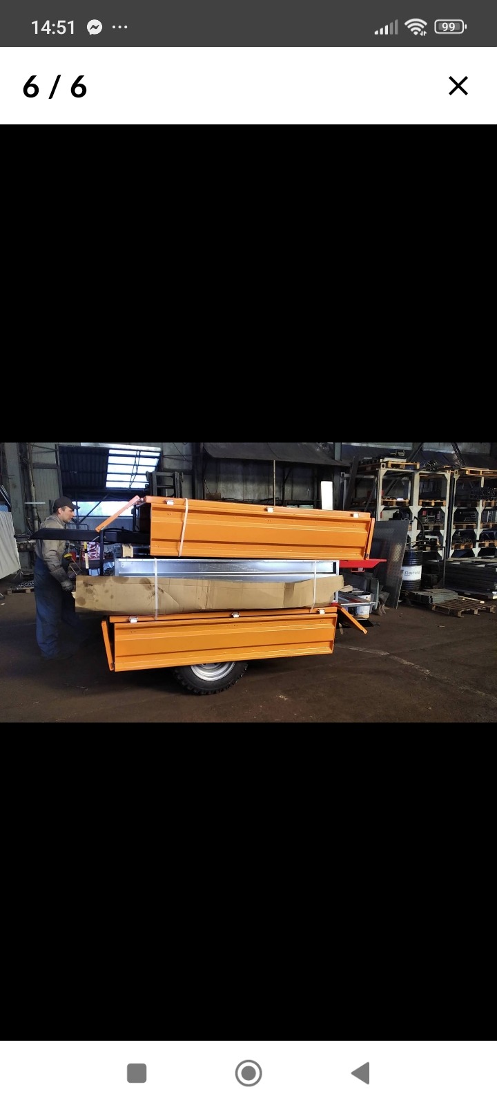 Pracownik w kombinezonie zabezpiecza pomarańczowe skrzynie na przyczepie z pojedynczą osią, w tle widoczny warsztat z metalowymi regałami i oknami.