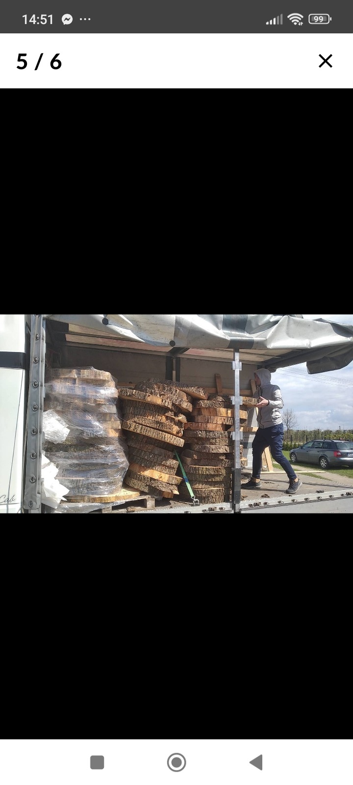 Wnętrze busa wypełnione okrągłymi plastrami drewna, część zapakowana w folię stretch, mężczyzna w bluzie z kapturem przenosi kolejny plaster, na zewnątrz widoczny samochód osobowy.