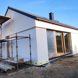 Se-bud - Nowoczesny, parterowy dom jednorodzinny z jasną elewacją w trakcie prac wykończeniowych, widoczne rusztowania i okna zabezpieczone folią, tło stanowi błękitne niebo i jesienne drzewa.