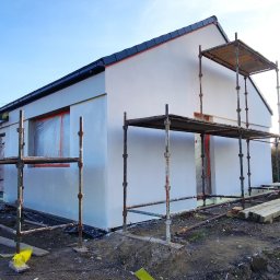 Se-bud - Nowo otynkowany, biały dom jednorodzinny w trakcie prac wykończeniowych elewacji, z widocznymi rusztowaniami i oknami zabezpieczonymi folią malarską, na pierwszym planie pracownik miesza farbę.