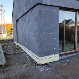 Se-bud - Narożnik domu jednorodzinnego w trakcie ocieplania elewacji, widoczne płyty styropianowe w kolorze grafitowym z białymi kołkami mocującymi, żółta izolacja przy gruncie i nowoczesne okno z drewnianą...