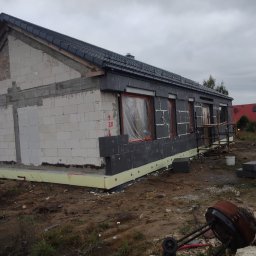 Se-bud - Budynek jednorodzinny w trakcie ocieplania elewacji zewnętrznej styropianem grafitowym, widoczne okna zabezpieczone folią, na ziemi gruz i narzędzia budowlane, pochmurne niebo w tle.