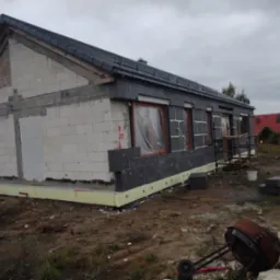 Budynek jednorodzinny w trakcie ocieplania elewacji zewnętrznej styropianem grafitowym, widoczne okna zabezpieczone folią, na ziemi gruz i narzędzia budowlane, pochmurne niebo w tle.