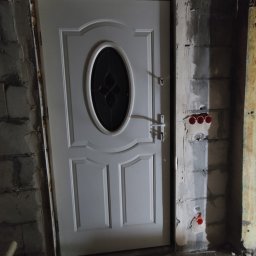 Se-bud - Białe drzwi wejściowe z owalnym przeszkleniem, osadzone w surowym otworze ściennym z widocznymi blokami betonowymi i instalacją elektryczną z czerwonymi rurkami, w trakcie prac wykończeniowych.