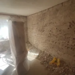 Pokój w trakcie skuwania tynku ze ścian, widoczne narzędzia na stole, zabezpieczone folią drzwi i gruz na podłodze.