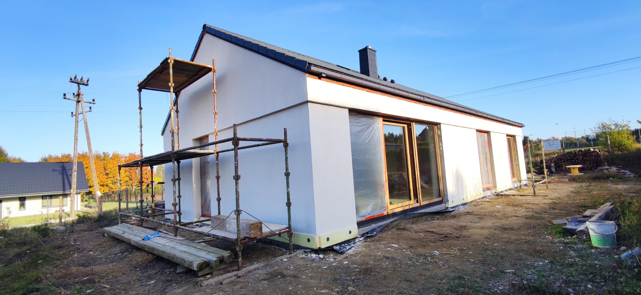 Nowoczesny, parterowy dom jednorodzinny z jasną elewacją w trakcie prac wykończeniowych, widoczne rusztowania i okna zabezpieczone folią, tło stanowi błękitne niebo i jesienne drzewa.