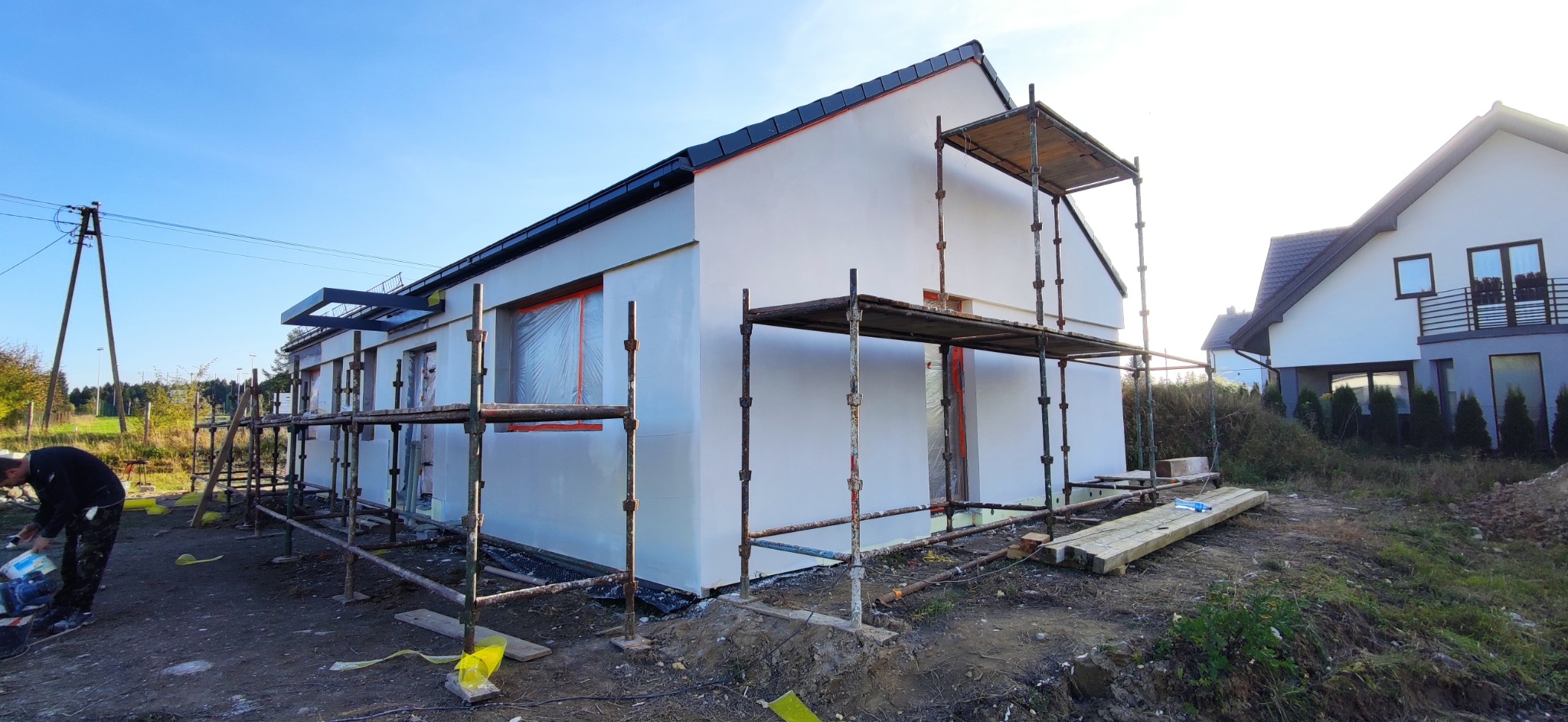 Nowo otynkowany, biały dom jednorodzinny w trakcie prac wykończeniowych elewacji, z widocznymi rusztowaniami i oknami zabezpieczonymi folią malarską, na pierwszym planie pracownik miesza farbę.