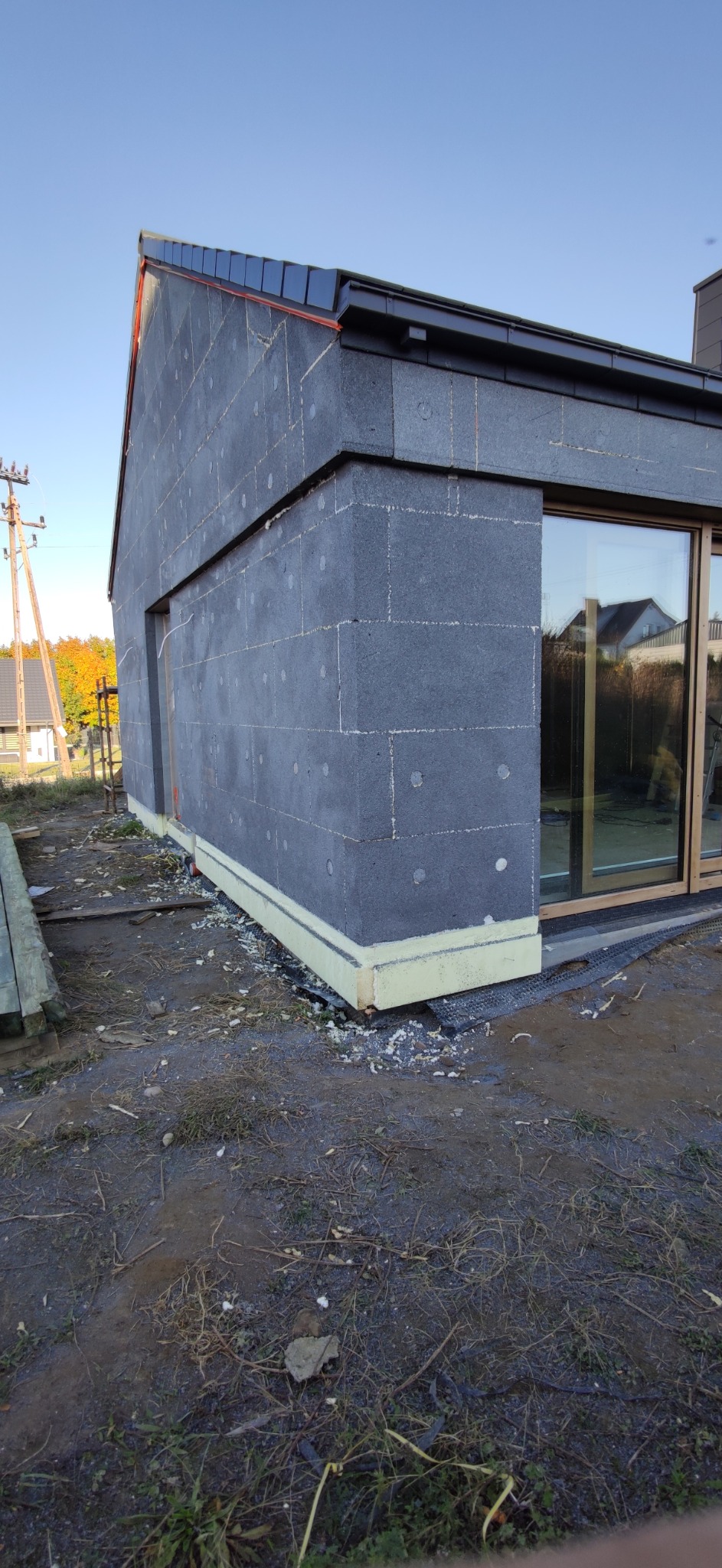 Narożnik domu jednorodzinnego w trakcie ocieplania elewacji, widoczne płyty styropianowe w kolorze grafitowym z białymi kołkami mocującymi, żółta izolacja przy gruncie i nowoczesne okno z drewnianą...