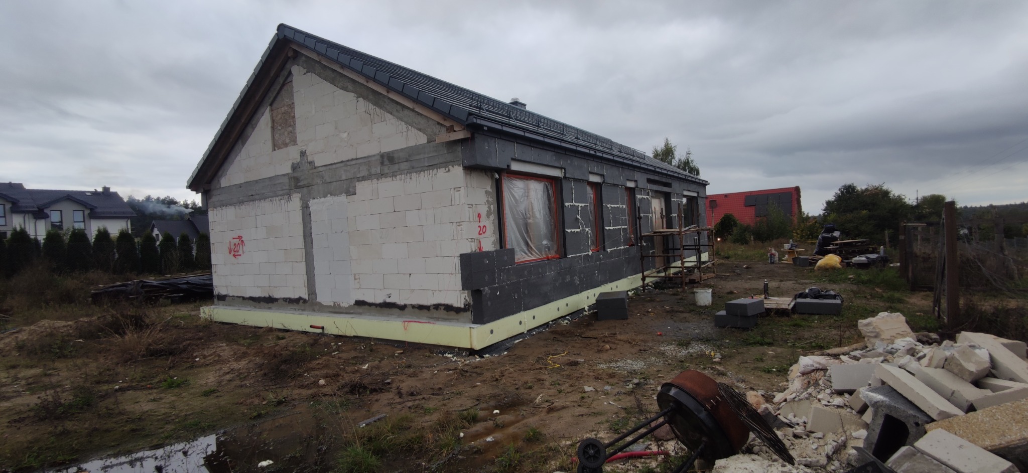 Budynek jednorodzinny w trakcie ocieplania elewacji zewnętrznej styropianem grafitowym, widoczne okna zabezpieczone folią, na ziemi gruz i narzędzia budowlane, pochmurne niebo w tle.