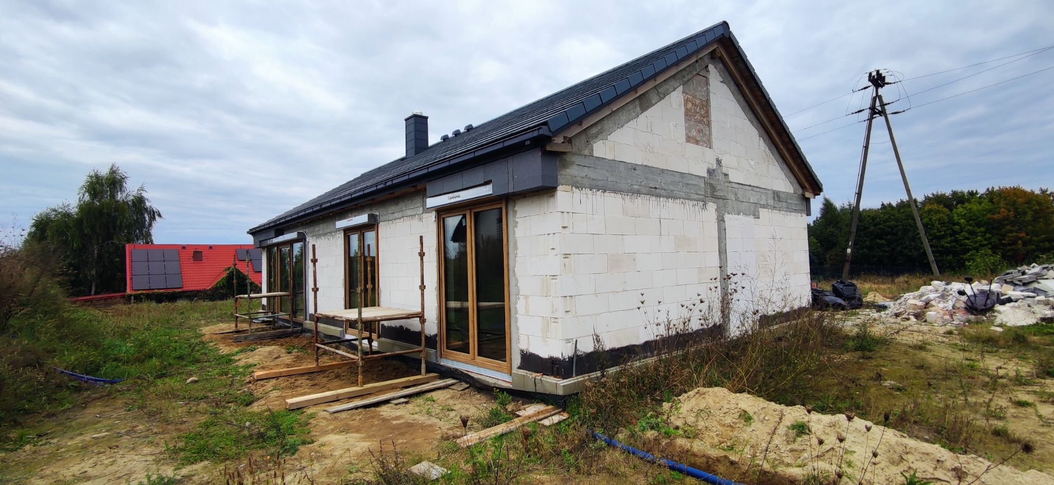 Budynek jednorodzinny w trakcie budowy, ściany z białego bloczka, drewniane okna, czarny dach, widoczne rusztowania i materiały budowlane na zewnątrz, w tle dom z panelami słonecznymi na dachu.