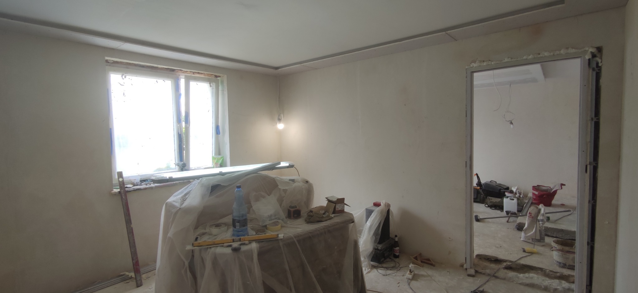 Wnętrze w trakcie prac wykończeniowych: ściany po nałożeniu gładzi, zabezpieczone okno, rozstawione narzędzia i materiały budowlane, widoczna sufitowa listwa dekoracyjna.