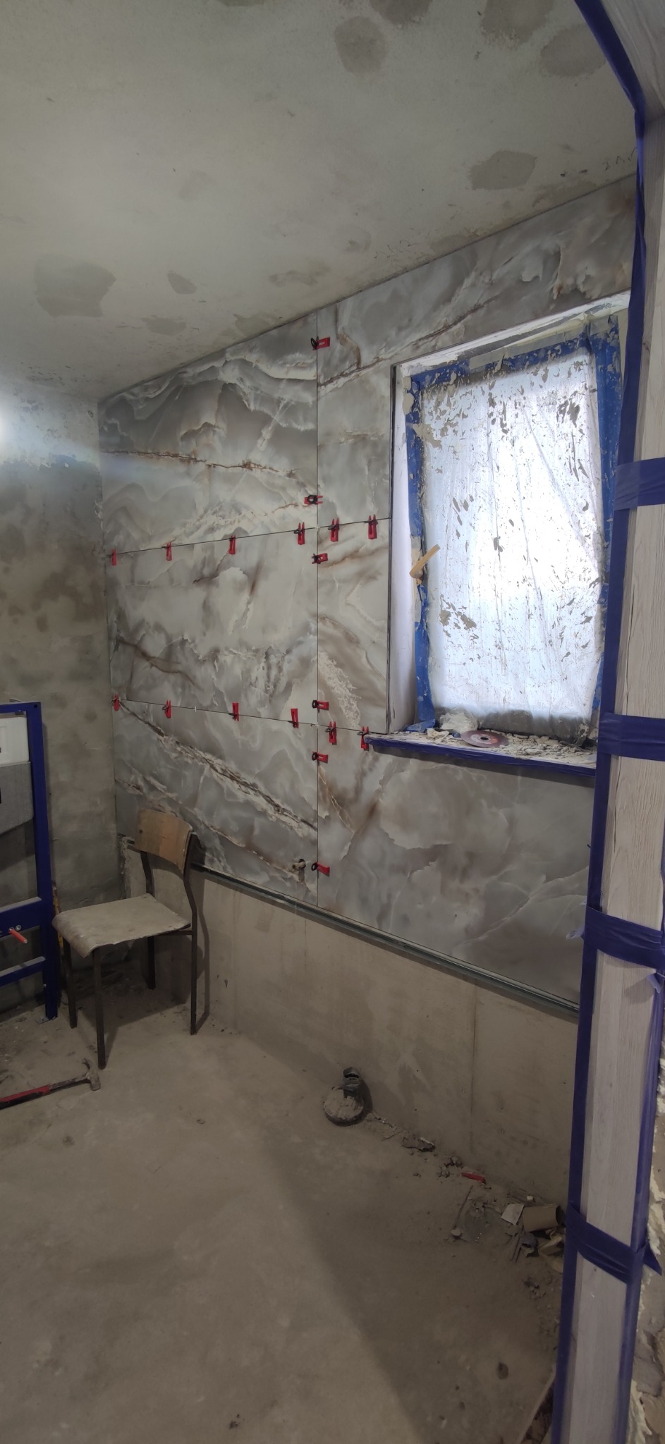 Montaż płytek imitujących marmur na ścianie w łazience, widoczne czerwone krzyżyki dystansowe, okno zabezpieczone folią, surowy stan podłogi i ścian.