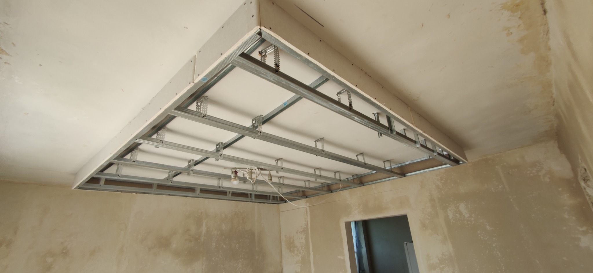 Montaż sufitu podwieszanego z płyt kartonowo-gipsowych na metalowej konstrukcji w surowym wnętrzu przed wykończeniem, widoczne przewody oświetleniowe.