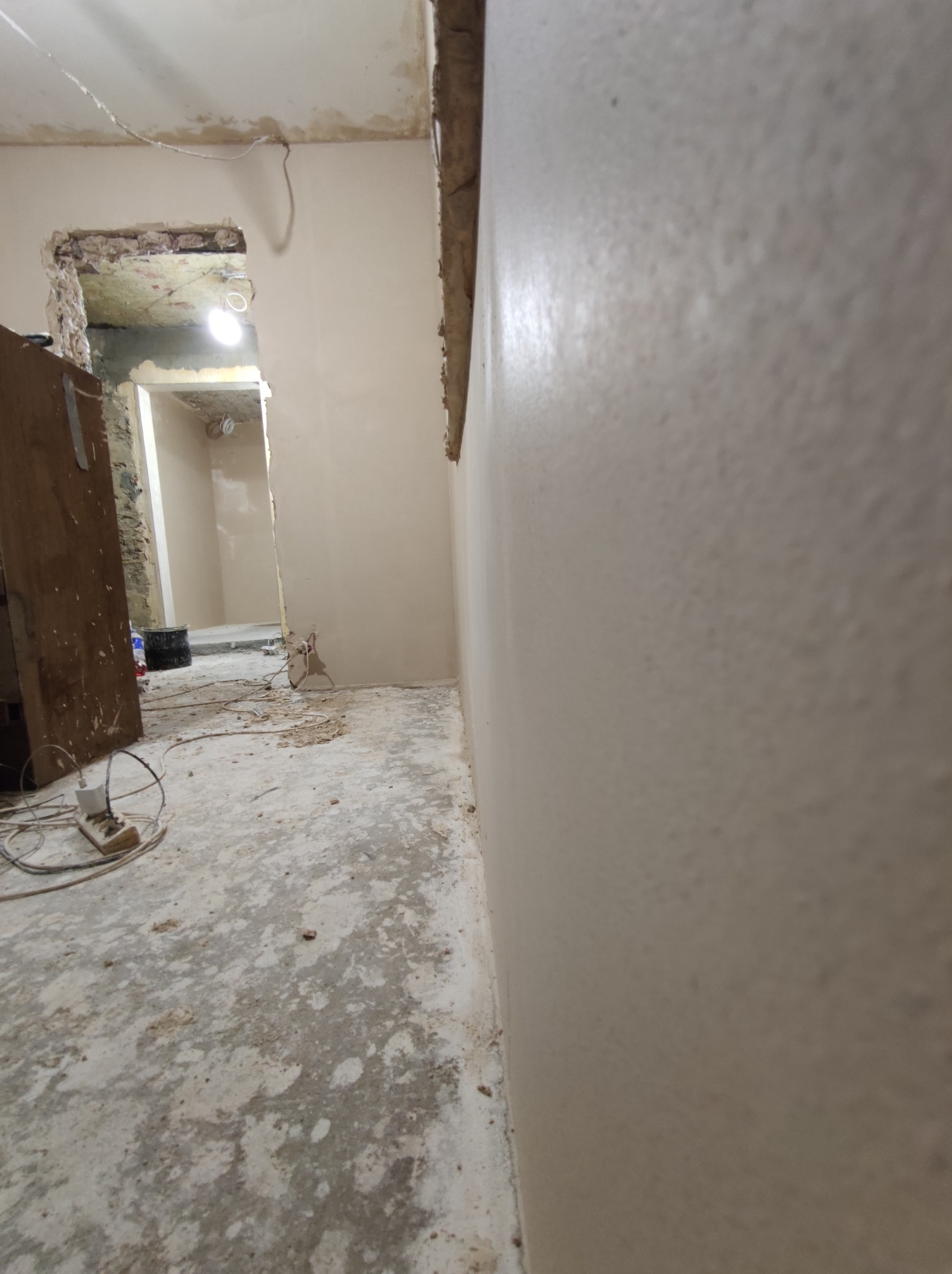 Świeżo wygładzona i pomalowana ściana w trakcie remontu, widoczne kable, przedłużacz i surowy beton na podłodze. W tle widoczne przejście do kolejnego pomieszczenia w stanie surowym.